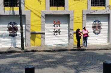 Two women standing in front of a yellow building with colorful skulls painted on the shuttered doors and windows.