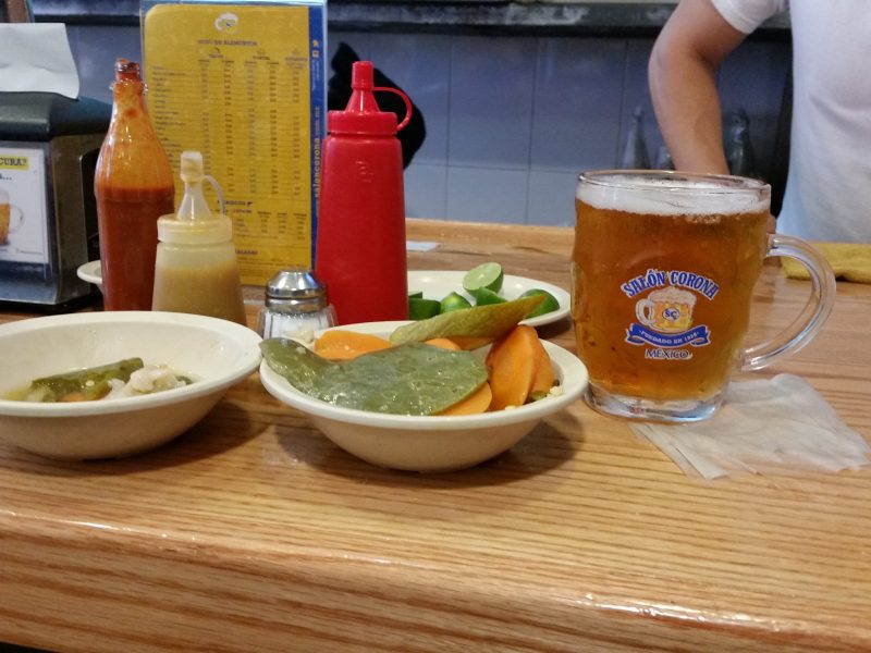 A bowl of pickled vegetables and a glass mug of beer sitting on a wooden counter with some condiment bottles in the background.