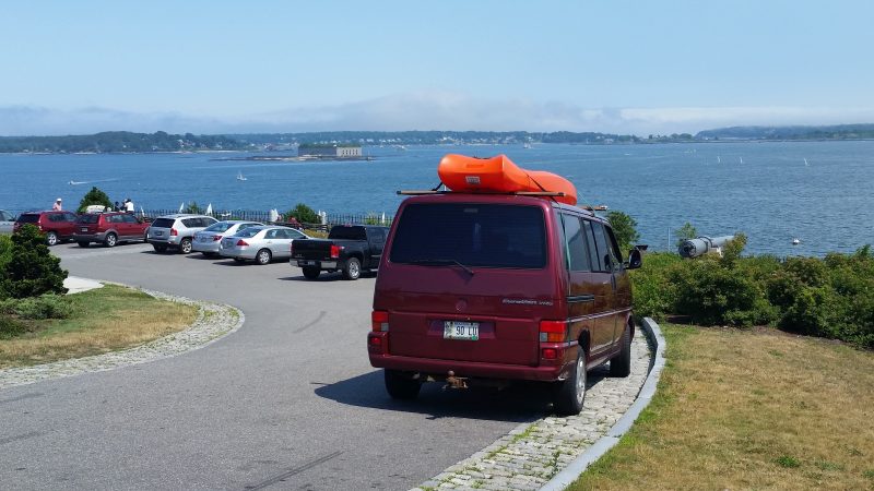 Burgundy Volkswagen van with an orange kayak on top of it and a bay in the background.