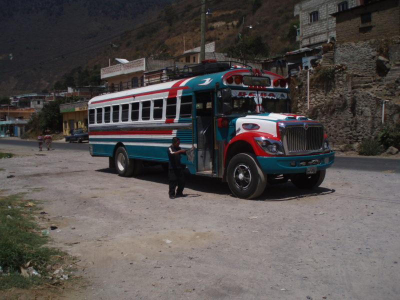 Man holding a rag standing next to a brightly-colored chicken bus in Zunil, Guatemala.
