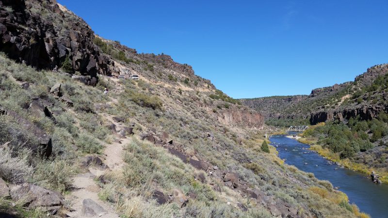 A path alongside a river with a bridge in the background.