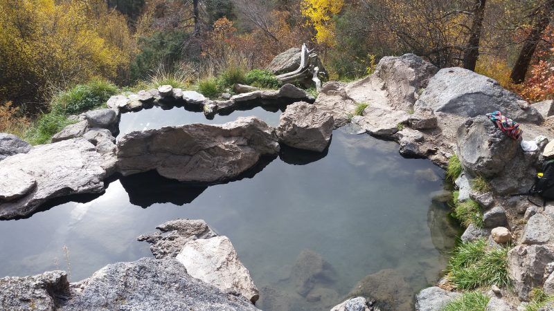 A pool at Spence Hot Springs filled with clear hot water reflecting the sun with pine trees in the background.