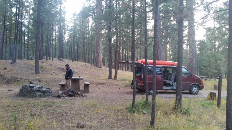 A burgundy van backed up to camping spot with a man standing outside of it looking at a campfire.