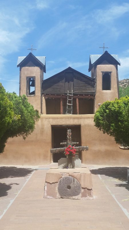 An old adobe church with two tress in front along the High Road to Taos from Santa Fe.