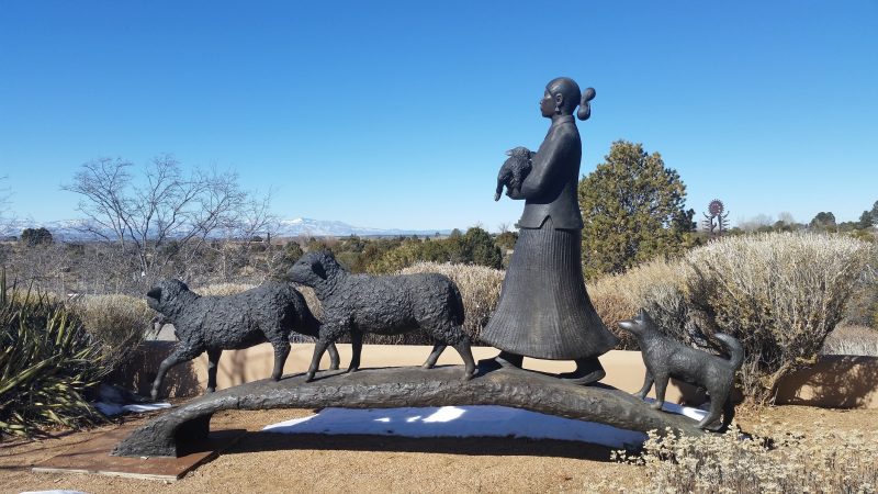 A bronze statue at Museum Hill in Santa Fe depicting a woman walking with two sheep and a small dog.