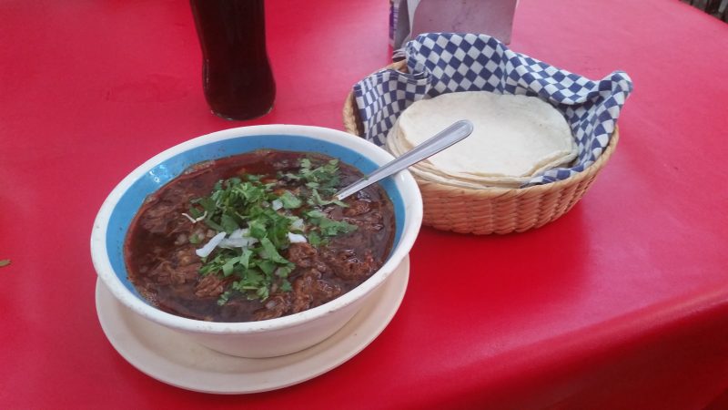 A bowl of birria stew on a red table, accompanied by a basket of corn tortillas.