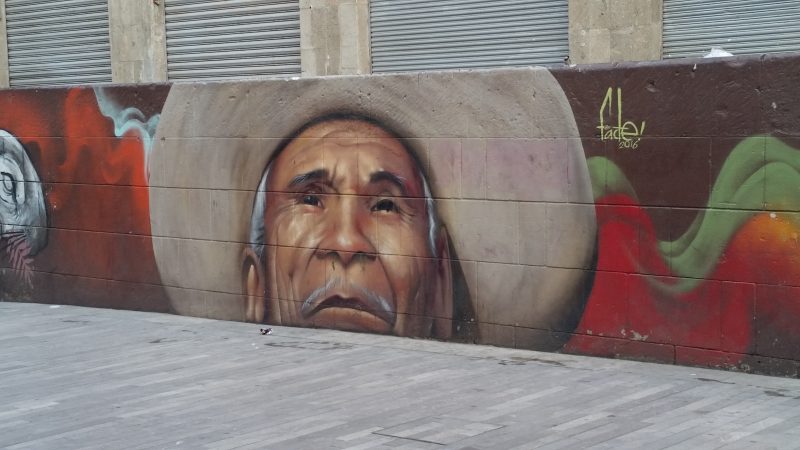 A pedestrianized area in Mexico City featuring a street art painting of an old man wearing a hat.