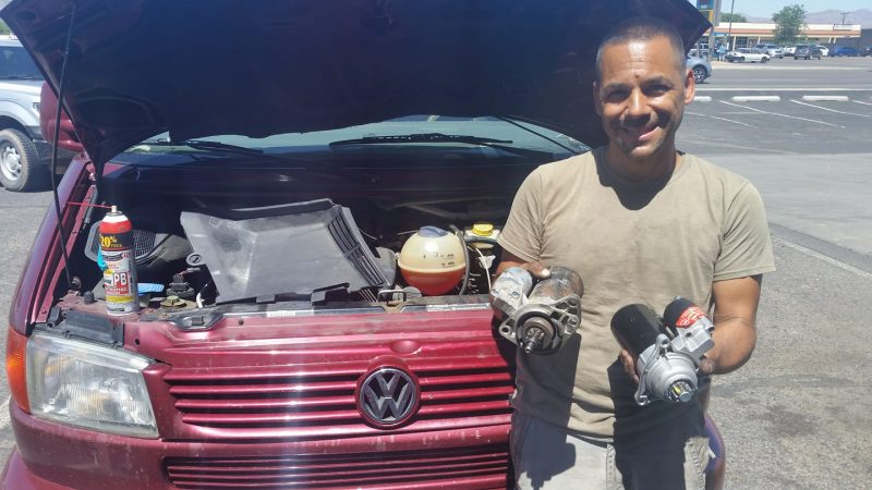 A man holding two starter motors in front of burgundy Volkswagen van with the hood up.