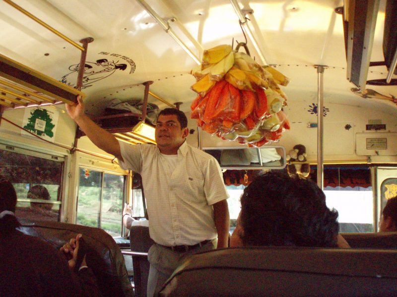 A man in a white shirt selling goods while standing in the front of a chicken bus in Guatemala.