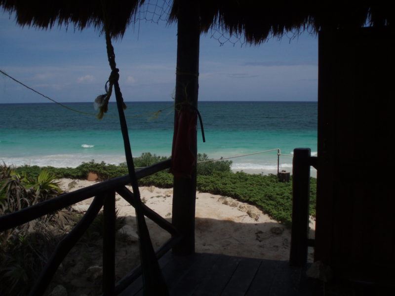 A porch with palapa roof with a light blue ocean in the background.